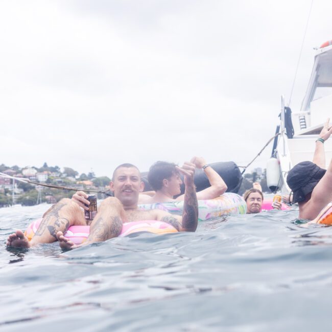 A group of people relax and enjoy drinks on inflatable pool floats in the water, next to a boat. The scene appears festive and laid-back, with cloudy skies in the background.