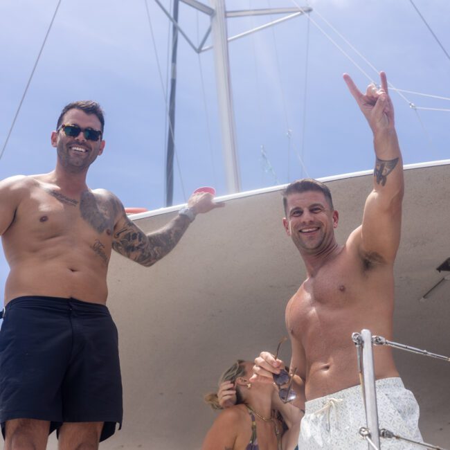 Two men on a boat, both shirtless and smiling. One is holding up a peace sign. A woman is partially visible behind them. It's a sunny day with a clear blue sky.