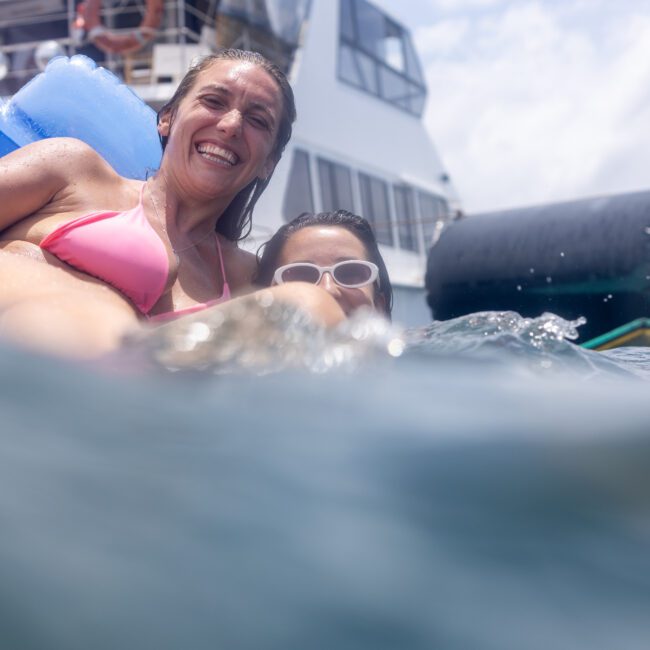 A woman in a pink bikini smiles while floating on a blue inflatable in the water. Another person, wearing sunglasses, is partially visible beside her. A yacht and a cloudy sky are in the background.