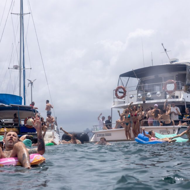 People relax and float on inflatables in the water near two anchored boats. One boat is crowded with people enjoying the sunny day. The scene is lively, with cloudy skies in the background.