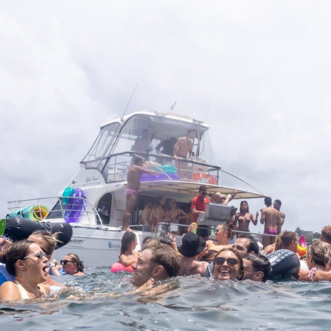 A crowd of people enjoying a party in the water near anchored yachts. Many are on inflatable pool floats and appear to be having a good time under a cloudy sky.