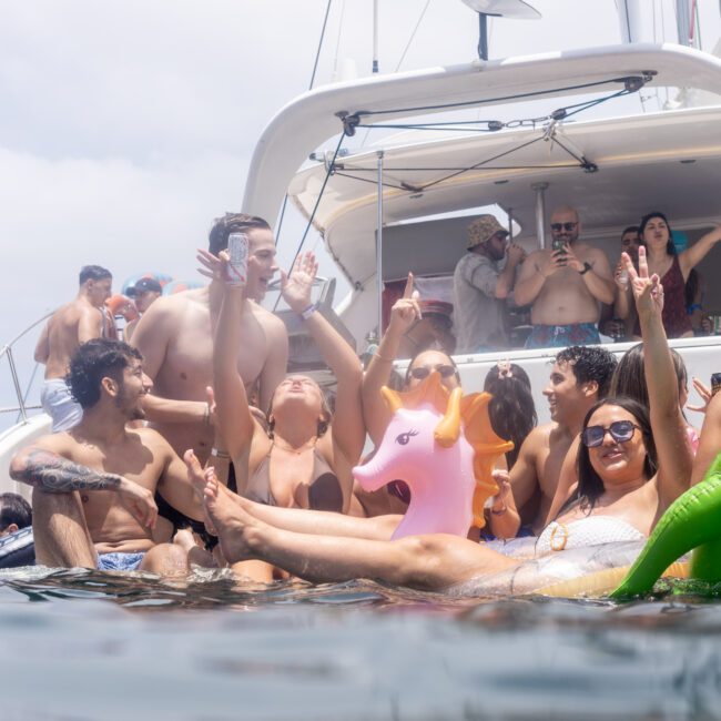 A group of people in swimsuits are enjoying a party on a yacht. Some are cheering with their arms up, while others relax in the water with a pink inflatable unicorn. The sky is cloudy, and there are more people on the yacht.