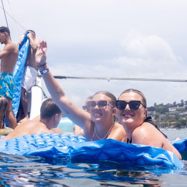 A group of people enjoys a sunny day on the water, waving from a blue inflatable raft. Some are relaxing on a boat nearby. They appear to be having fun in an idyllic setting with hills in the background.