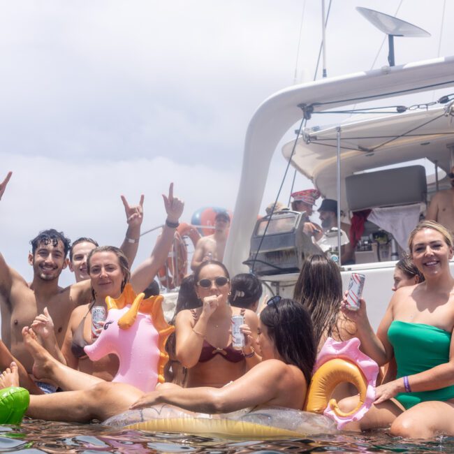 A group of people enjoying a party on a boat, some in the water, wearing swimwear, and holding inflatable toys shaped like animals. They are smiling, posing, and making hand gestures. The boat's deck and a cloudy sky are visible in the background.