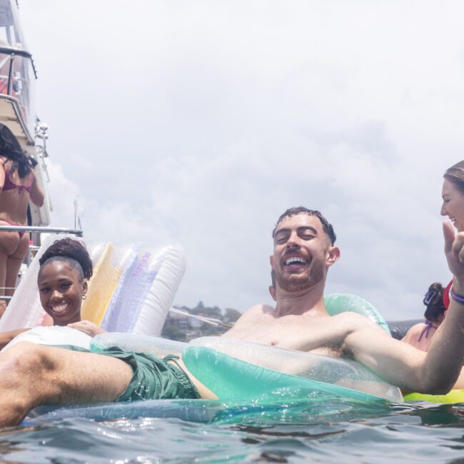 A group of people relaxes on inflatable rafts in the water near a boat. A man in the foreground smiles and gives a thumbs up. The sky is cloudy, and everyone appears to be enjoying the sunny, fun atmosphere.