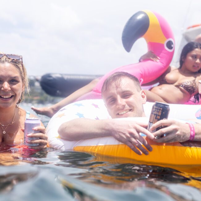 A group of people enjoying a sunny day in the water. Two people are in the foreground, smiling while floating on inflatable pool toys, including a pink flamingo. Others are relaxing on a boat in the background.