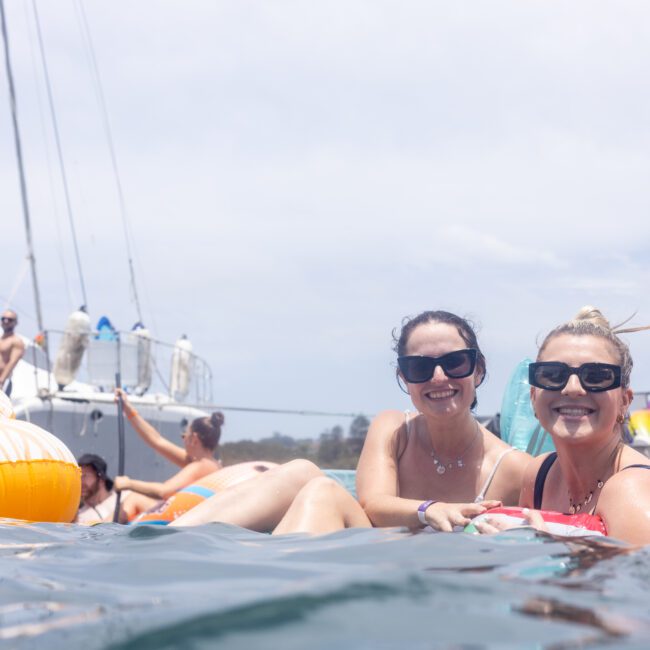 Two people smile at the camera while floating in the water on inflatable rings. They are near a boat with others in the background, enjoying a sunny day.