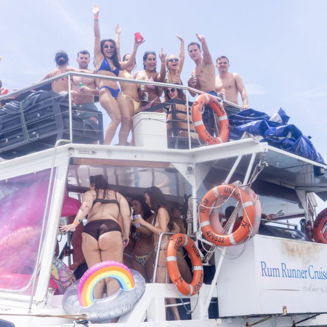 A group of people in swimwear are enjoying a sunny day on a boat named "Rum Runner Cruises." Some are standing on the upper deck waving, while others are below. Lifebuoys and an inflatable rainbow float are visible. The sky is clear and blue.