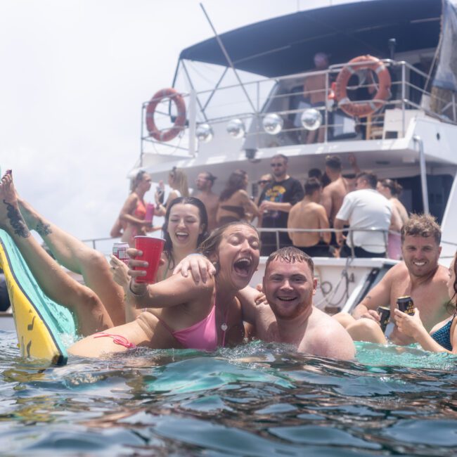 A group of people laughing and enjoying themselves on floating mats in the water near a boat. They are holding drinks and appear to be having a fun time at a party with other guests on the vessel in the background.