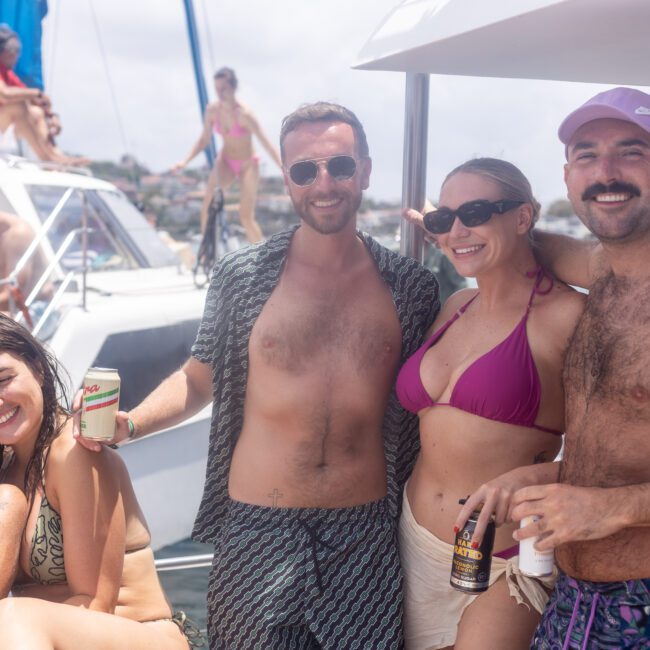 A group of people enjoying a sunny day on a boat. They are smiling and holding drinks, wearing swimsuits and sunglasses. The boat is on the water, with a clear sky and distant land visible in the background.