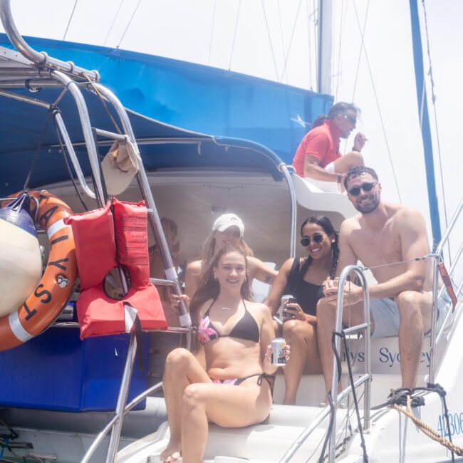 A group of people relax and smile on a sailboat under a bright sky. They sit on the steps, some holding drinks. The boat has visible lifebuoys and a sunshade. Everyone is dressed in swimwear, enjoying a sunny day on the water.