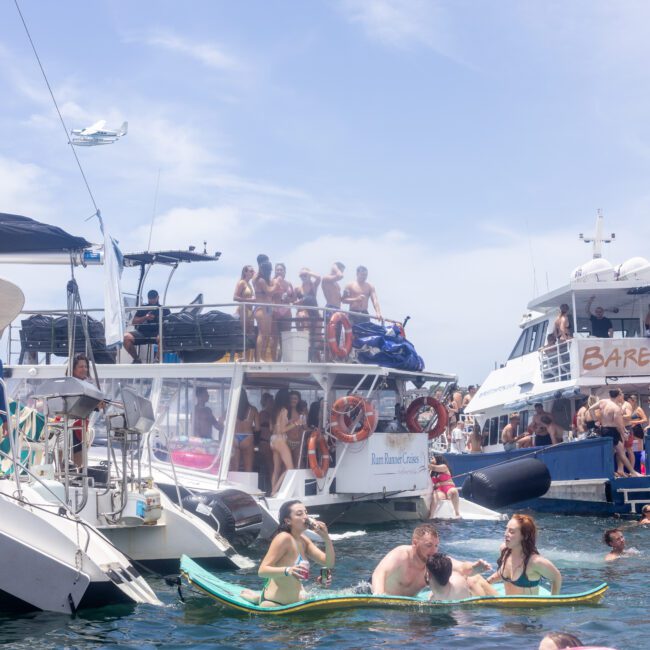 A lively scene with people enjoying a sunny day on boats and in the water. Groups are gathered on decks and inflatables. A plane flies overhead, and the boat on the right has a "Barefoot Blue" sign.