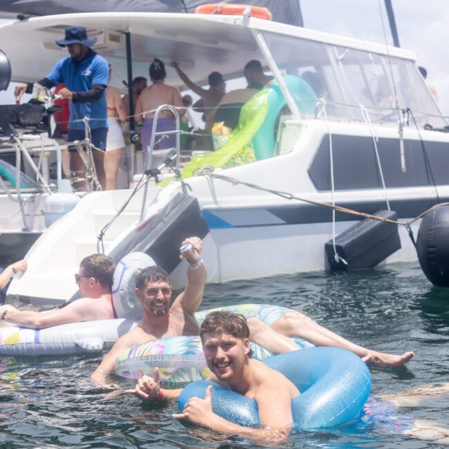 A group of people enjoys a sunny day on the water, lounging on inflatable tubes next to a boat. Some are floating in the water while others relax onboard. A man gives a thumbs-up gesture, and a grill is visible on the boat.