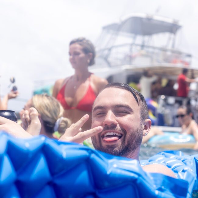 A man with a beard enjoys floating on a blue raft, making a peace sign and smiling. In the background, several people relax on a boat under a bright, sunny sky.