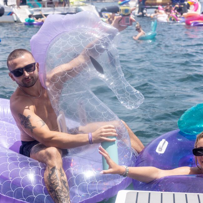 Two people relax on inflatable pool toys in a lake. The man sits on a purple flamingo with a clear seahorse float, and the woman on a purple float reaches up with a drink. A group of people and more floats are in the background on the water.