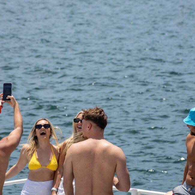 A group of young adults enjoying a sunny day on a boat, with the ocean in the background. One person is taking a selfie, while others laugh and hold drinks.