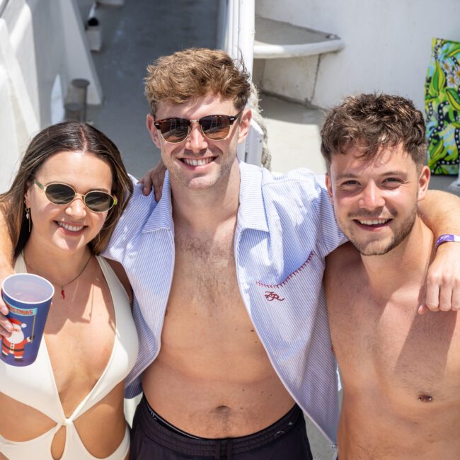 Three people smiling on a boat deck. One woman and two men are wearing swimsuits and sunglasses, with the men having their arms around each other's shoulders. They're standing in sunny weather.