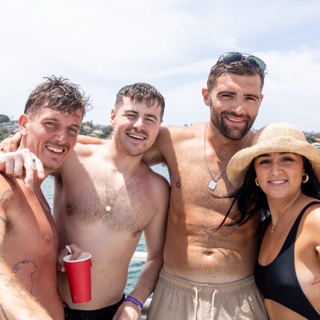 Four people smiling and enjoying a sunny day on a boat, holding drinks. The background features a cityscape by the water. One person is wearing a sun hat and sunglasses.