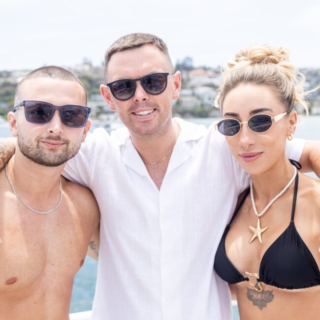 Three people on a boat, all wearing sunglasses. The person in the middle has a white shirt, and the individuals on either side are in swimsuits. They are posing with arms around each other, with a scenic waterfront backdrop.