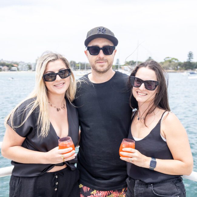 Three people stand together on a boat, smiling at the camera. Two women hold orange drinks; one of them wears a black outfit, and the other a white top. The man in the middle wears sunglasses and a cap. A body of water and boats are visible in the background.