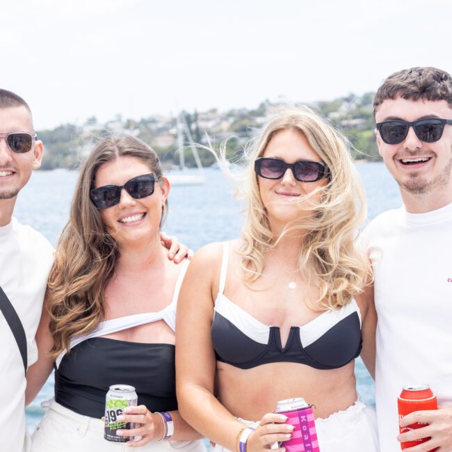 Four people wearing sunglasses, smiling and holding canned drinks, pose together outdoors. A body of water with boats is visible in the background, along with green hills and a cloudy sky.