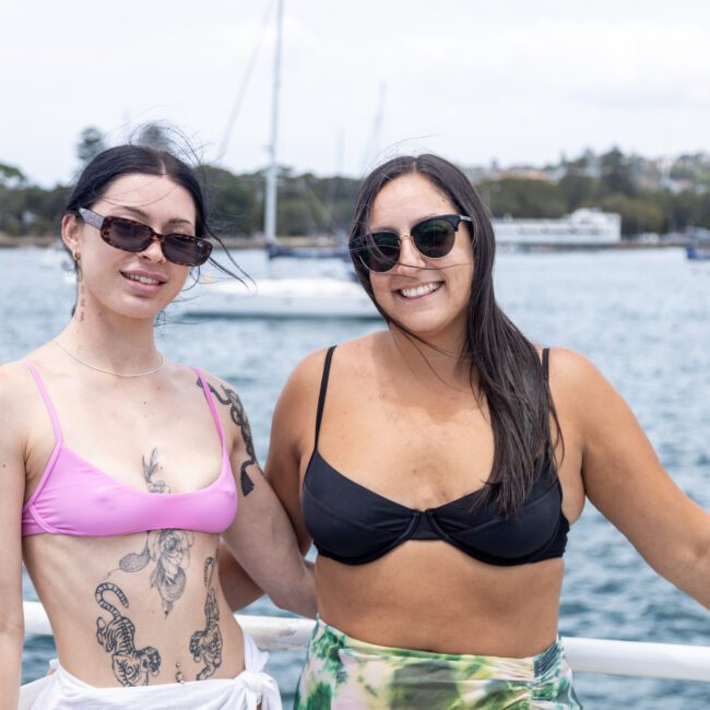 Two women in sunglasses, one with a pink bikini top and tattoos, and the other with a black bikini top, standing on a boat. A scenic view of water, a sailboat, and distant shoreline in the background under a cloudy sky.