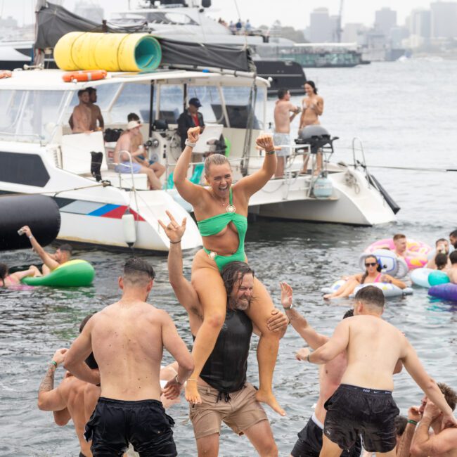 A group of people enjoy a party on a floating platform. A woman in a green bikini rides on a man's shoulders. Boats and inflatables are visible in the background under a cloudy sky.