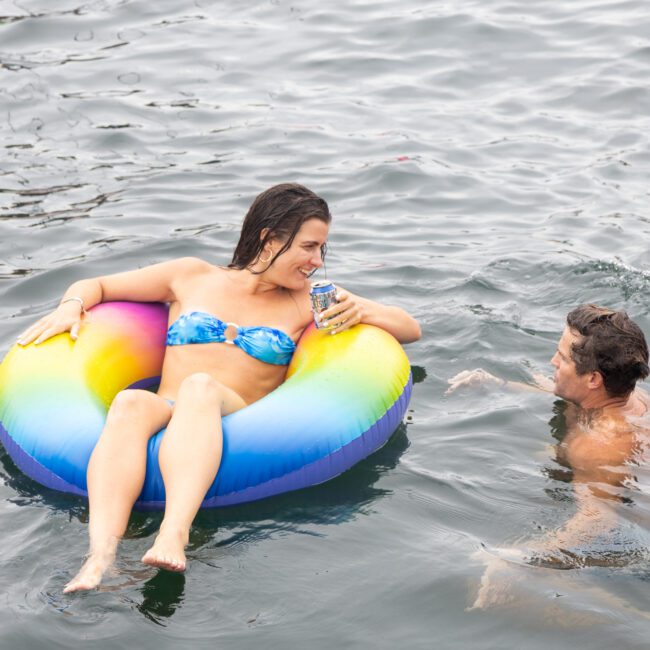 A woman in a blue bikini is lounging on a rainbow-colored inflatable ring, holding a drink. A man is swimming next to her in the water. Both are smiling and enjoying the moment in the open water.