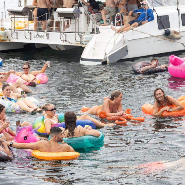 People are relaxing on colorful inflatable floats in the water near a boat. Some are swimming or sitting on the edge of the vessel, enjoying a lively and fun atmosphere on a sunny day.