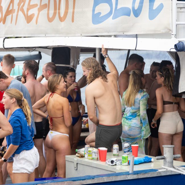 A group of people in swimsuits gather on a boat deck under a canopy that reads "Barefoot Blue." They are socializing and enjoying drinks. The scene appears lively and relaxed, set against a backdrop of water and trees.