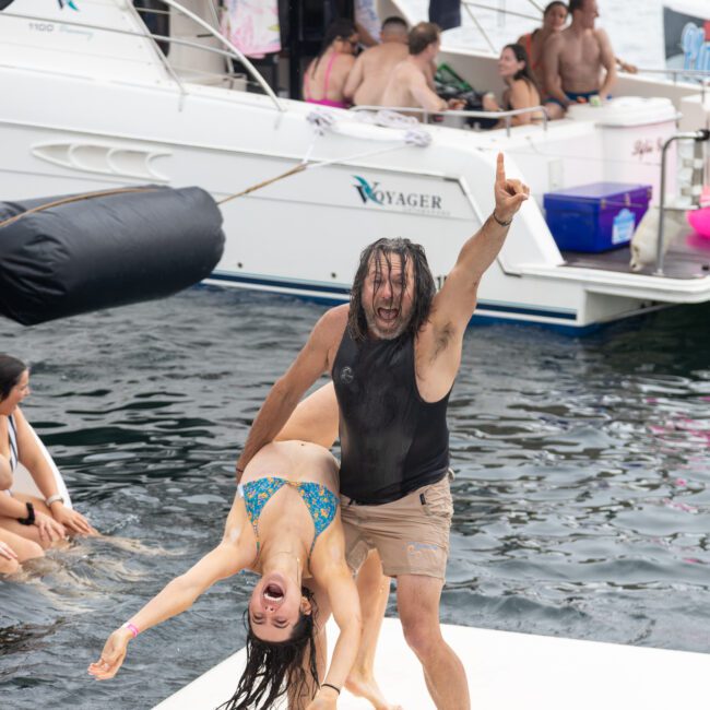 A joyful man and woman dance energetically on a platform over water. The woman is in a backbend, and the man points upwards. People relax on a nearby boat, adding to the festive atmosphere.