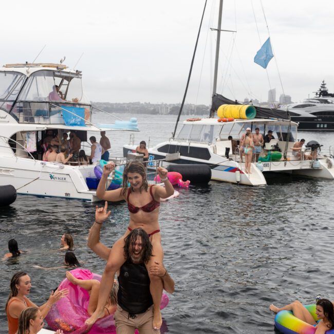 A lively yacht party with people swimming and having fun. A man carries a woman on his shoulders while others float on inflatables near boats. Overcast sky and cityscape in the background create a festive and relaxed atmosphere.