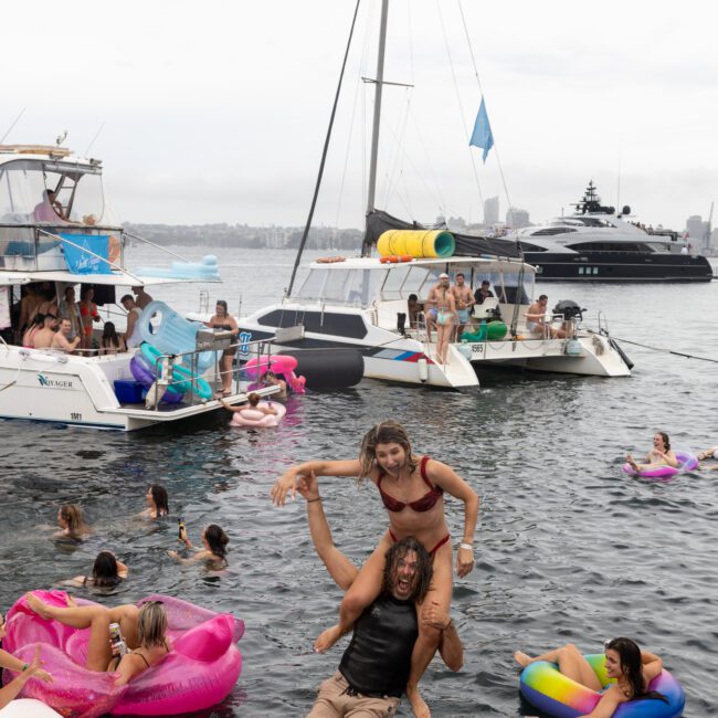 A festive scene on the water with people enjoying a boat party. A man carries a woman on his shoulders in the foreground, surrounded by others relaxing on colorful inflatable floats. Several boats and yachts are anchored in the background.