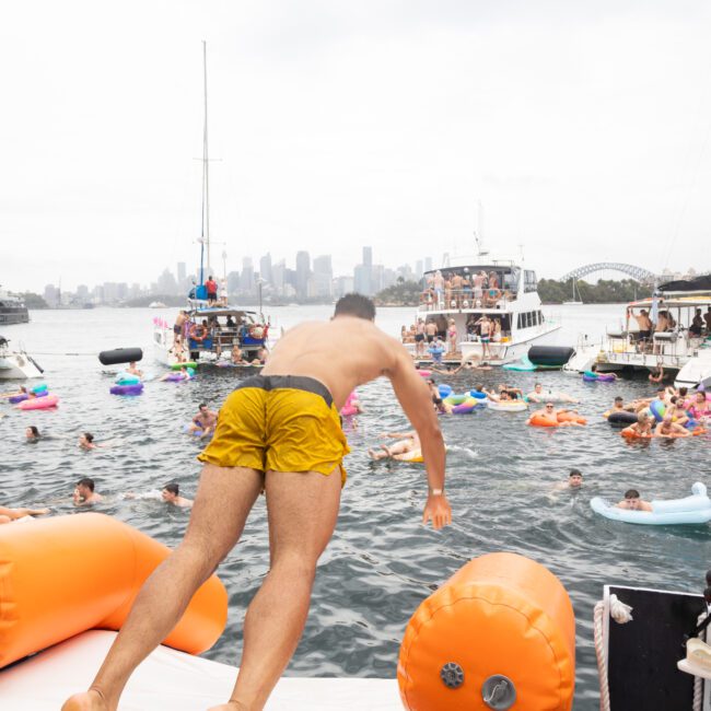 A person in yellow shorts dives from a boat into a crowded harbor filled with colorful inflatables and people swimming. Several boats are anchored nearby with the city skyline in the background under a cloudy sky.