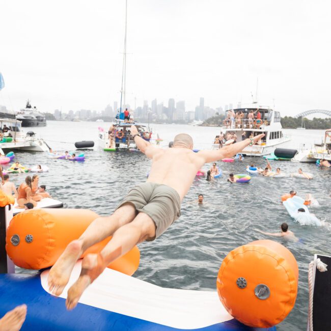 A man is diving from a float into a crowded bay, surrounded by boats and people swimming. The background features a city skyline and overcast sky. Others on floats and a woman in a yellow shirt watch nearby.