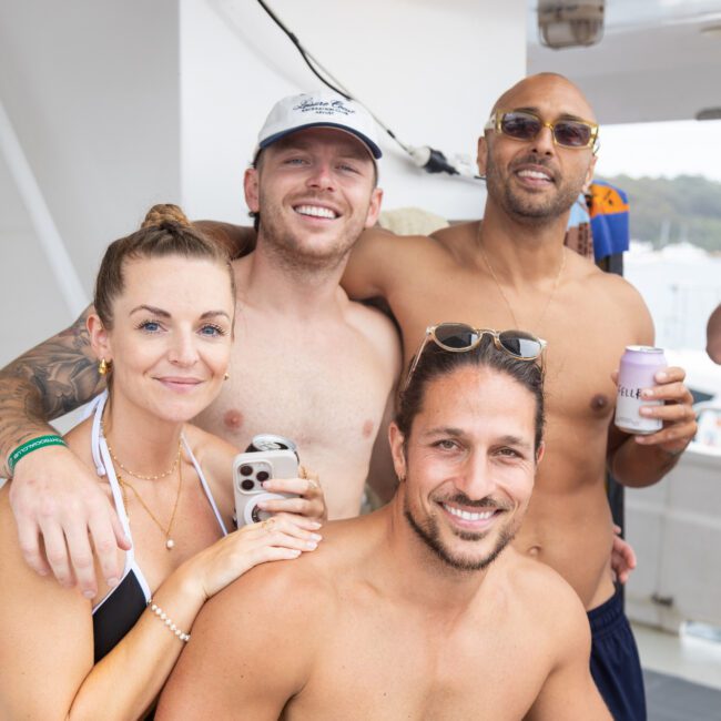 A group of four people, three men and one woman, smiling and posing on a boat. Two are holding drinks. They are in swimwear, and the background shows part of the boat and a blurred view of the water and shoreline.