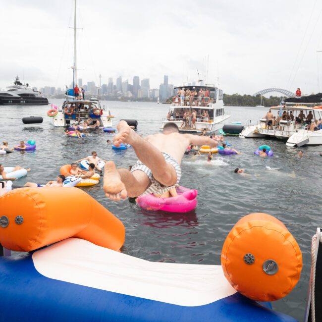 A man jumps into the water from a boat, surrounded by others on colorful inflatable devices. Several boats are anchored nearby, and a city skyline with a bridge is visible in the background. The scene is lively and festive.