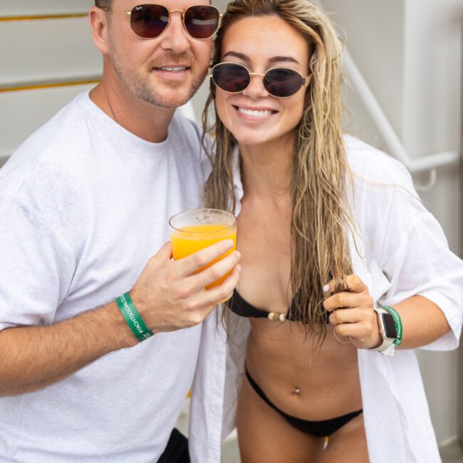 A man and woman in sunglasses smile at the camera on a boat. The man wears a white t-shirt and holds a drink. The woman is in a bikini and open shirt, holding a drink. Both have green wristbands. Stairs are visible in the background.