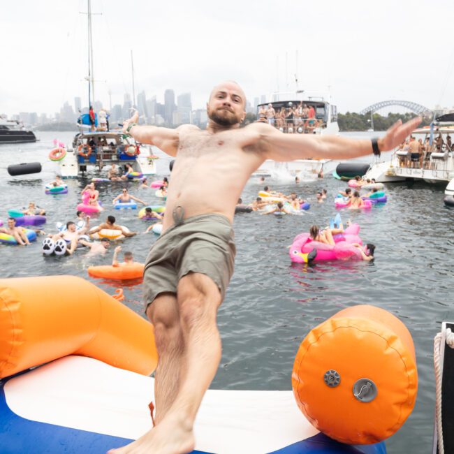 A shirtless man with shorts leans back playfully on an inflatable platform on a boat. Behind him, numerous people float on colorful inflatable rings in a marina, surrounded by boats and a city's skyline in the background.