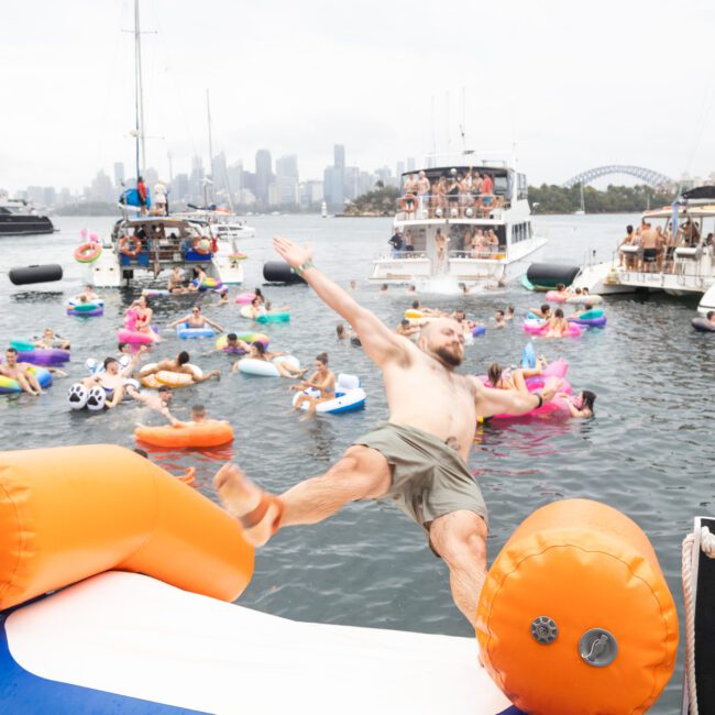 A man is captured mid-jump from an inflatable slide into a body of water. Numerous people are on boats and inflatables in the background, enjoying a lively party scene. The sky is overcast, and a city skyline is visible in the distance.