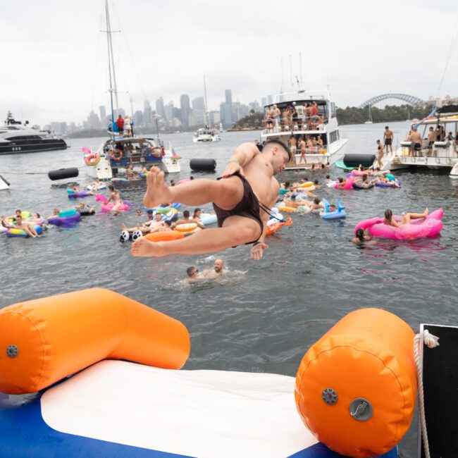 A person leaps from an inflatable platform into a bay filled with colorful inflatable floats. People in swimsuits enjoy the water, with boats and a city skyline in the background. The atmosphere is lively and festive.