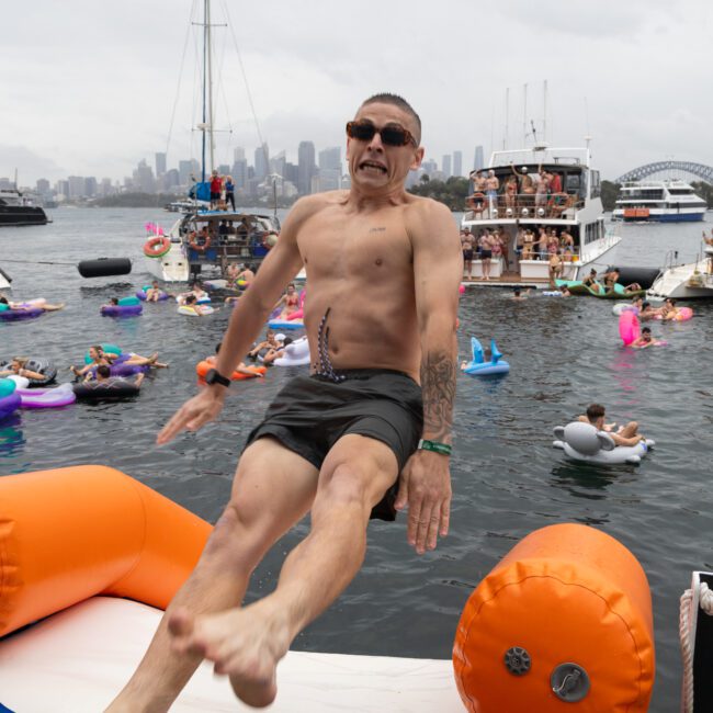 A man wearing sunglasses and shorts is sliding off an inflatable structure into a busy water scene with boats and people on floats. The background features a city skyline under a cloudy sky.