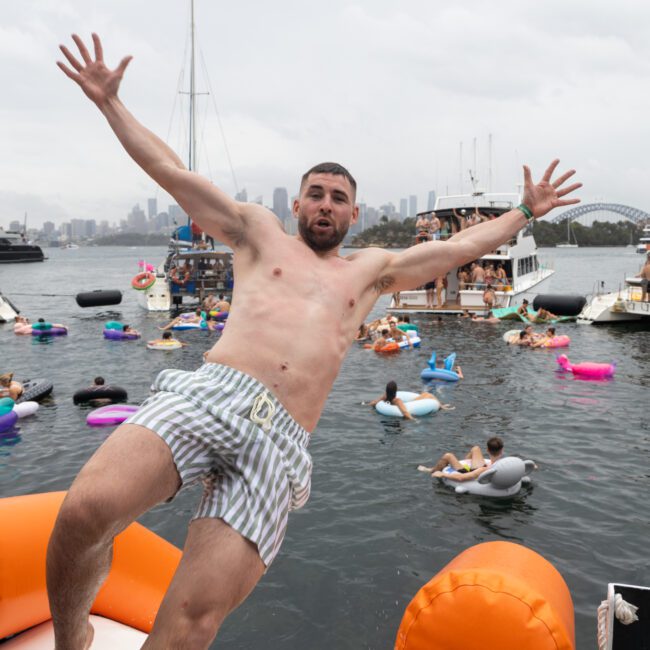 A man in striped swim trunks is mid-jump off an inflatable raft into the water, with arms outstretched. Behind him, numerous people are enjoying a party on boats and inflatable floats. The city skyline is visible in the background under a cloudy sky.