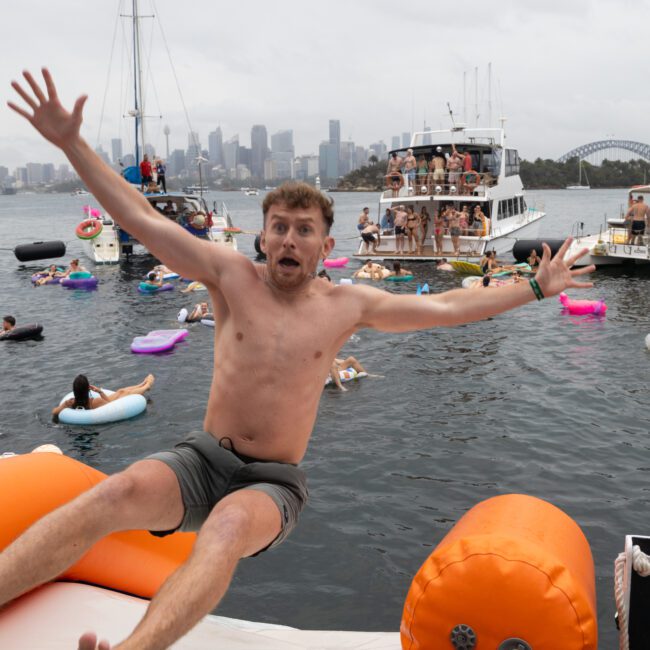 A man excitedly jumps from an inflatable platform into a body of water, with a backdrop of boats and people swimming. The scene suggests a festive atmosphere. Overcast sky and a city skyline, including a bridge, are visible in the distance.