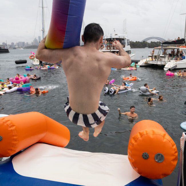 A man jumps from an inflatable structure into the water at a party with people on various colorful inflatables. Boats are anchored nearby, and a cityscape and bridge are visible in the background. The sky is overcast.