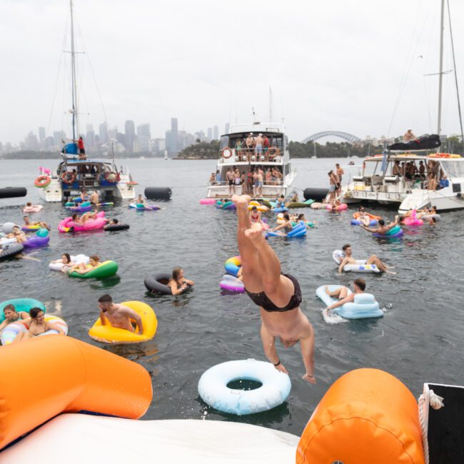 A person in swimwear is diving into the water from a colorful float. The background features boats and numerous people enjoying the water on inflatables, with a cityscape visible in the distance. The atmosphere is lively and festive.