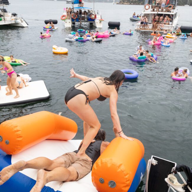 People are enjoying a floating party on a lake with colorful inflatables. A woman in a black bikini climbs onto inflatable cushions. Boats and more people are visible in the background, creating a lively and festive atmosphere.