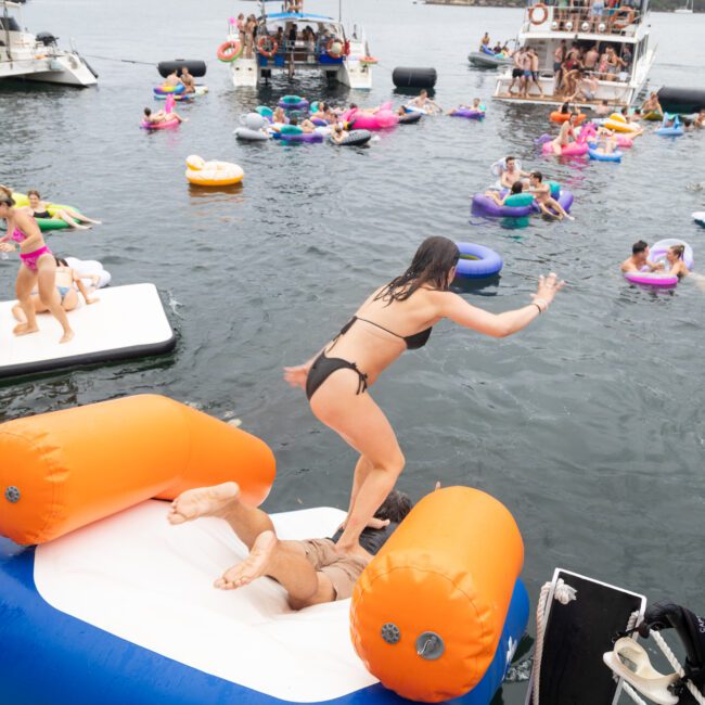 People are enjoying a day at the lake, lounging on colorful inflatable rafts and boats. A woman in a black swimsuit is jumping onto an inflatable slide with another person beneath her. The atmosphere is lively and festive.