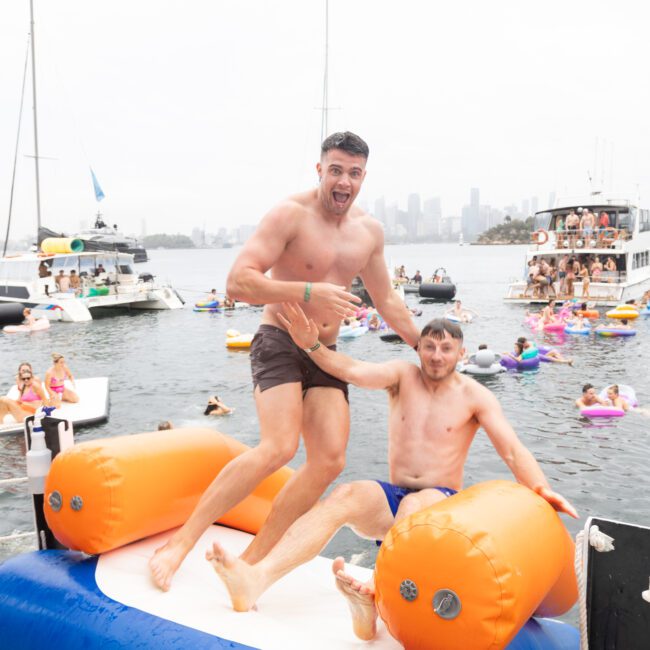 Two men are playfully wrestling on an inflatable platform in the water. Boats and people on floating rings are in the background, with a city skyline visible. The atmosphere is lively and fun.