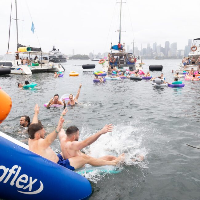 People enjoying a day on the water, with two individuals sliding off an inflatable into the sea. Boats and numerous people on inflatables fill the background, with city skyscrapers visible in the distance.